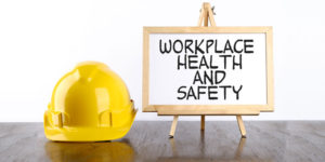 Picture of a yellow hard hat next to a small easel that is supporting a framed board displaying the words 'Workplace Health and Safety'.