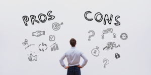 Man in light blue shirt with hands on hips staring at whiteboard with the words PROS and CONS displayed.