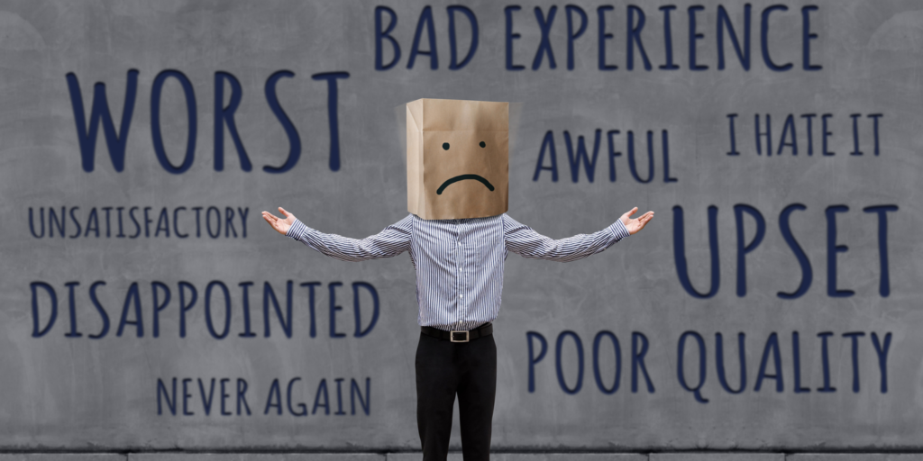 A businessman standing with arms raised to the side and with a brown paper bag over his head. The paper bag has an unhappy face drawn on it. The wall behind the man is full of words of complaint, e.g. worst, upset, disappointed.