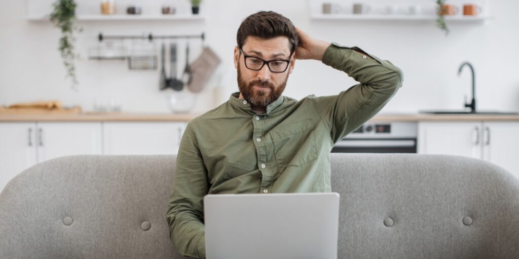 Man in his studio flat, sitting on the sofa, looking confused at his laptop.