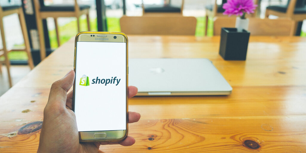 Male hand holding a smartphone with the Shopify logo displayed on the screen at an empty meeting table, illustrating the concept of the purge on meetings.