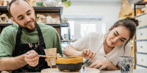 Cheerful partners in aprons colouring a clay bowl while working together in their ceramic workshop.