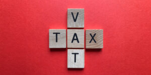 5 wooden blocks in a cross shape with 'TAX' written horizontally and 'VAT' written vertically, resting on a red background