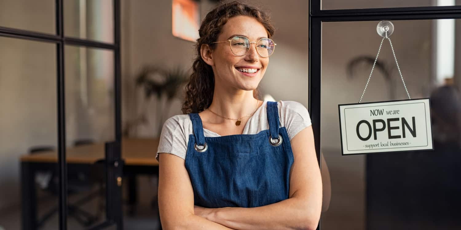 Portrait of smiling new business owner at her restaurant entrance with a sign 'Now we are OPEN' hanging beside the door.