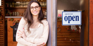 Portrait of smiling young female cafe owner woman standing with arms crossed in the doorway. Open sign on the glass door.