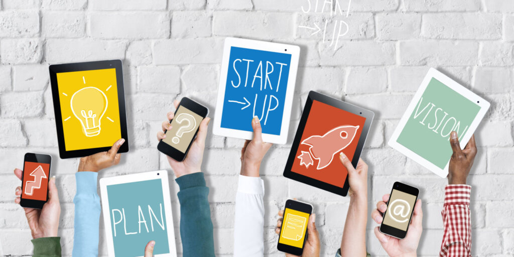 Group of hands holding up digital devices, against a white brick background, with Startup Concept messages displayed on the screens.