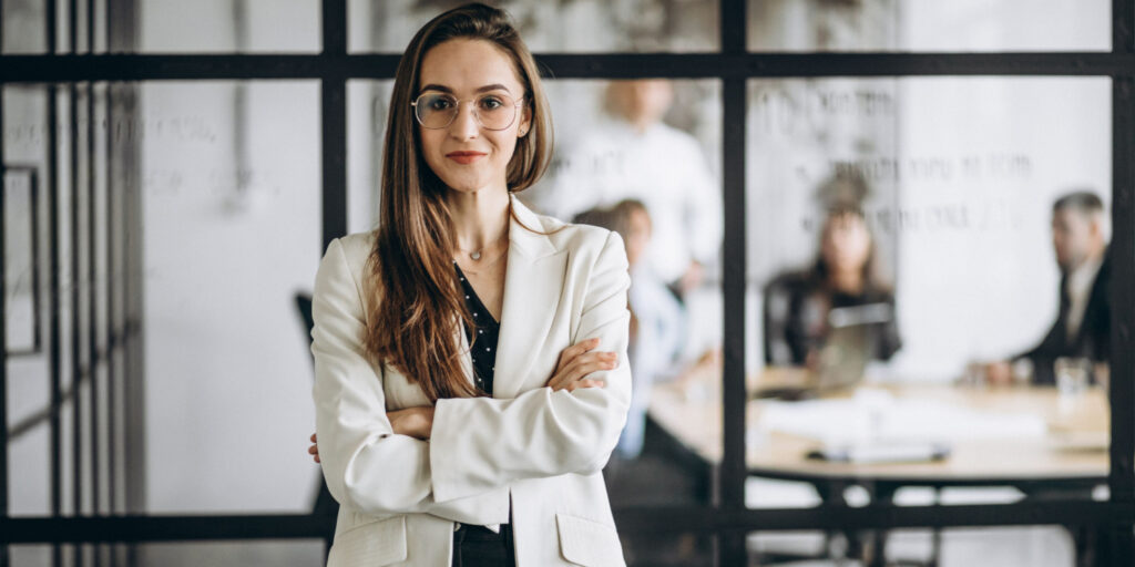 Portrait of a female company secretary standing in an office with a board meeting taking place behind her.