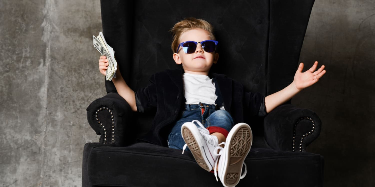 Happy young boy wearing sunglasses, sitting in a chair and holding a stack of bank notes.