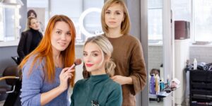 Beauty salon owner and assistant preparing a stylish makeup for a beautiful young blonde girl before start of a fashion show.