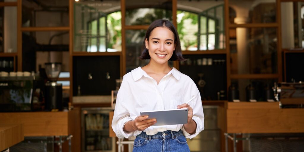 Portrait of an Asian female business owner standing with tablet in front of her cafe entrance. Concept of ethnic minority business in the UK.