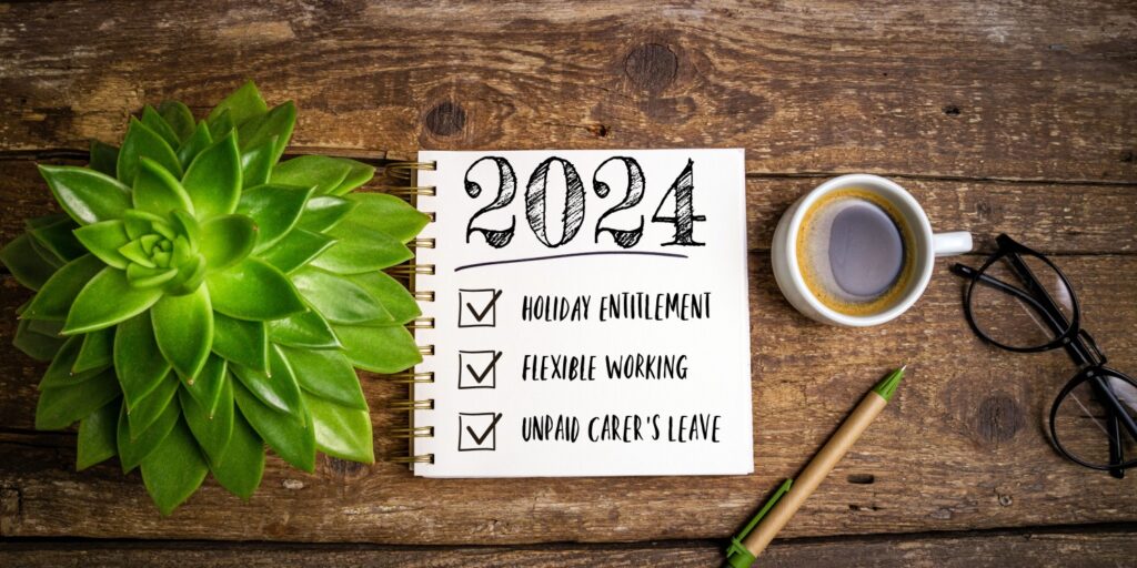 A notebook, coffee cup, and plant sitting on a wooden table. The notebook has a handwritten heading of '2024' with three tick points below: 'Holiday Entitlement', 'Flexible Working', and 'Unpaid Carer's Leave'.