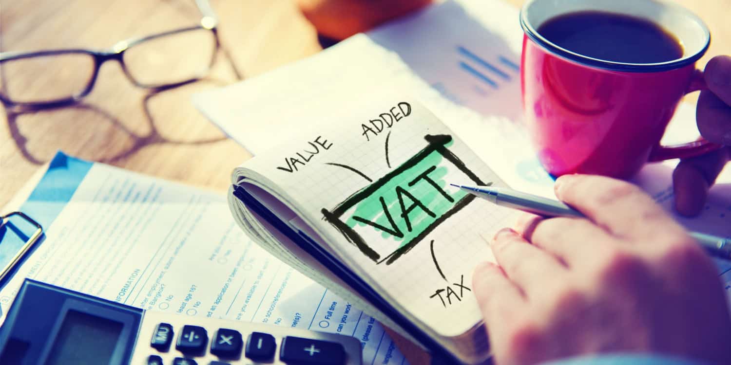 'VAT' and VALUE ADDED TAX' written on a notebook with a male hand holding a pen and a cup of coffee and pair of spectacles sitting in the background.