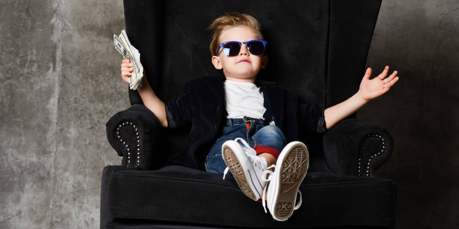 A happy young boy wearing sunglasses holding a stack of bank notes whilst sitting back in a large black armchair.