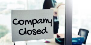 'Company closed' sign on the window of an empty office with businessman in the background packing his belongings and walking out of the business.