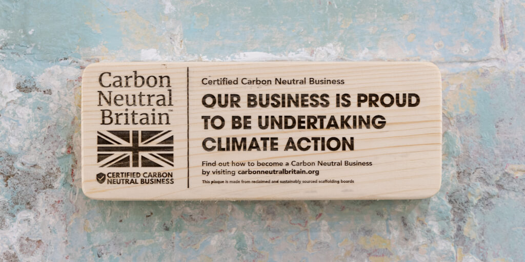 A wooden plaque on the wall of 1st Formations' reception which was provided by Carbon Neutral Britain and certifies our business's carbon neutral status. The main heading reads 'OUR BUSINESS IS PROUD TO BE UNTERTAKING CLIMATE ACTION.