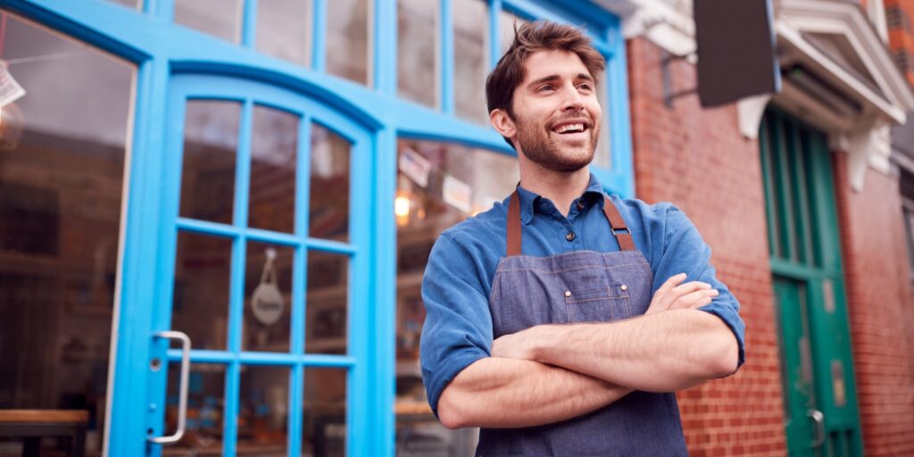 A handsome business owner standing in front of his shop's blue door on a UK high street. He's smiling because he's happier now than when he was an employee.