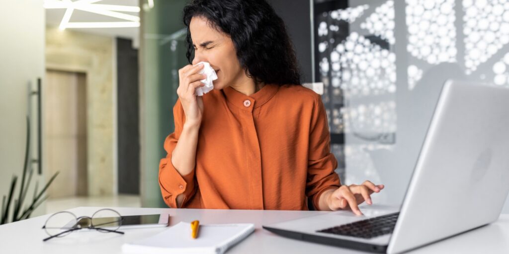 A young woman sitting at a desk in her office with laptop and sneezing, holding a handkerchief to her nose.