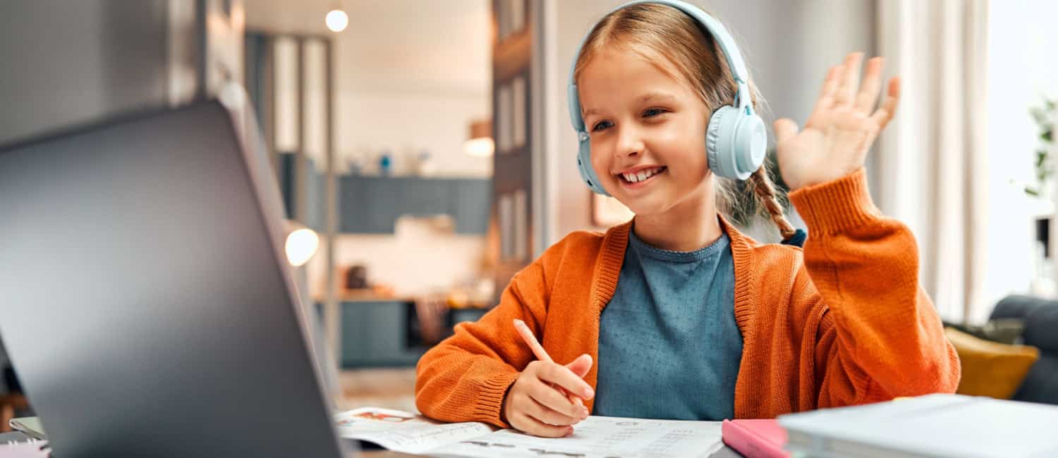 A smiling girl starting an online turorial wearing a headset and raising her hand with a greeting gesture while looking at a laptop screen.