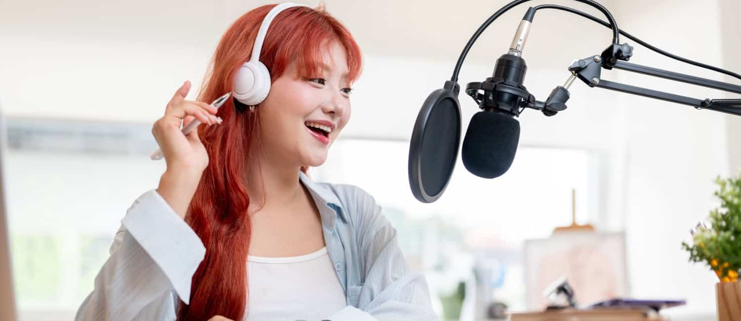 An attractive Asian female podcaster with long red hair recording her podcast in a studio, wearing headphones and speaking into a microphone.