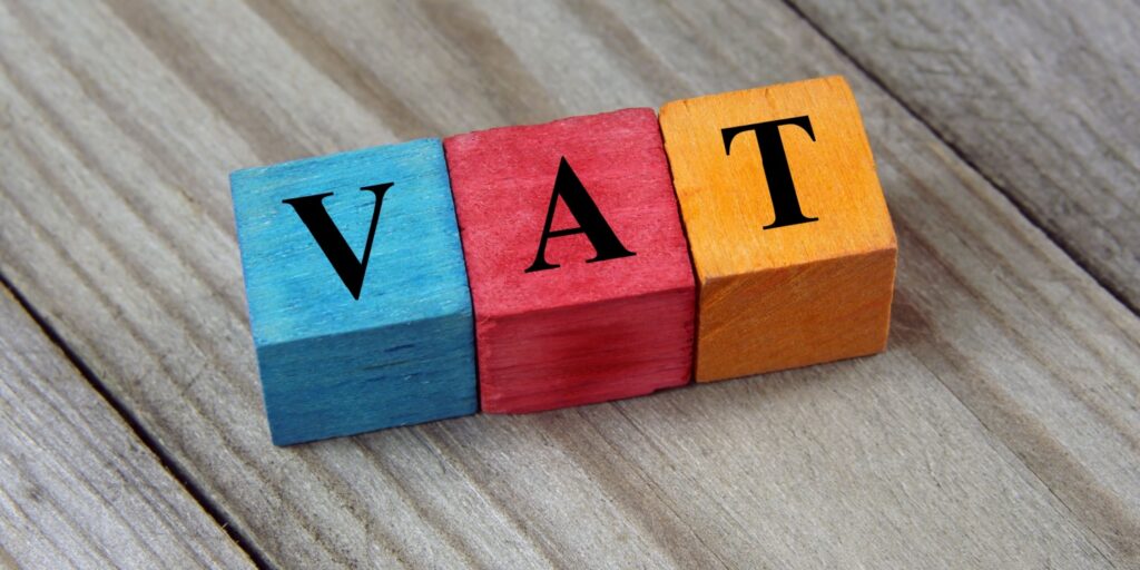 The text 'VAT' (Value Added Tax) printed in black font on three colourful wooden cubes lying on a wooden table.