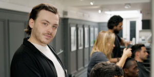 A portrait of a young male barber shop owner looking at the camera while cutting a client's hair with other barbers and customers in the background.