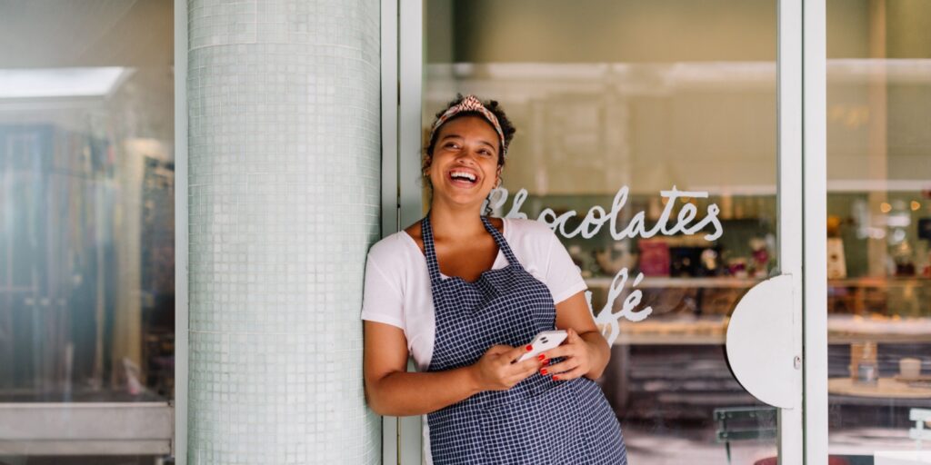 Small business owner leaning against column, holding a phone and smiling in front of their cafe, representing SME growth