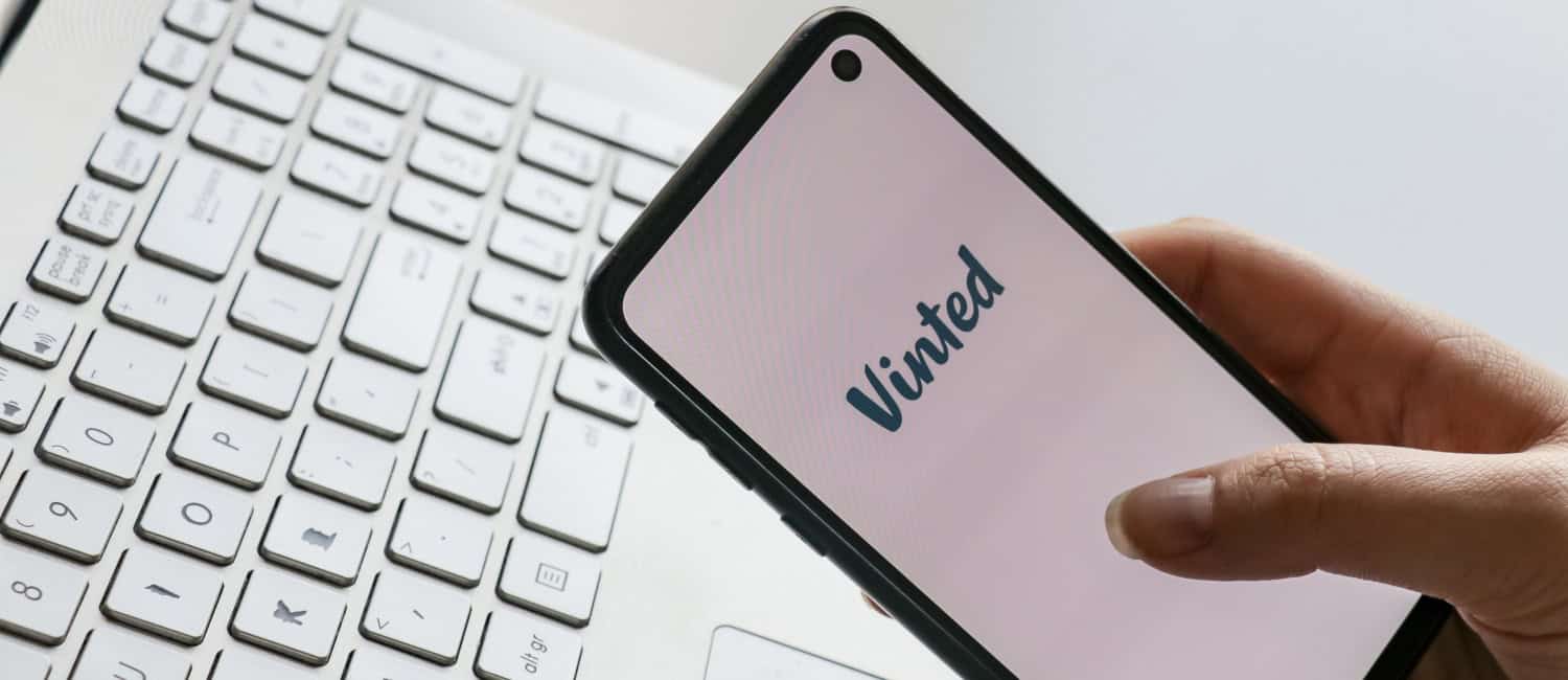 a hand holding a phone with the logo of the second hand selling app Vinted with a laptop in the background.