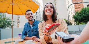 A customer happily pays for a coffee with her phone. The price strategy left her satisfied. She's sitting outside next to a man and she's smiling as she taps her phone to a card reader.