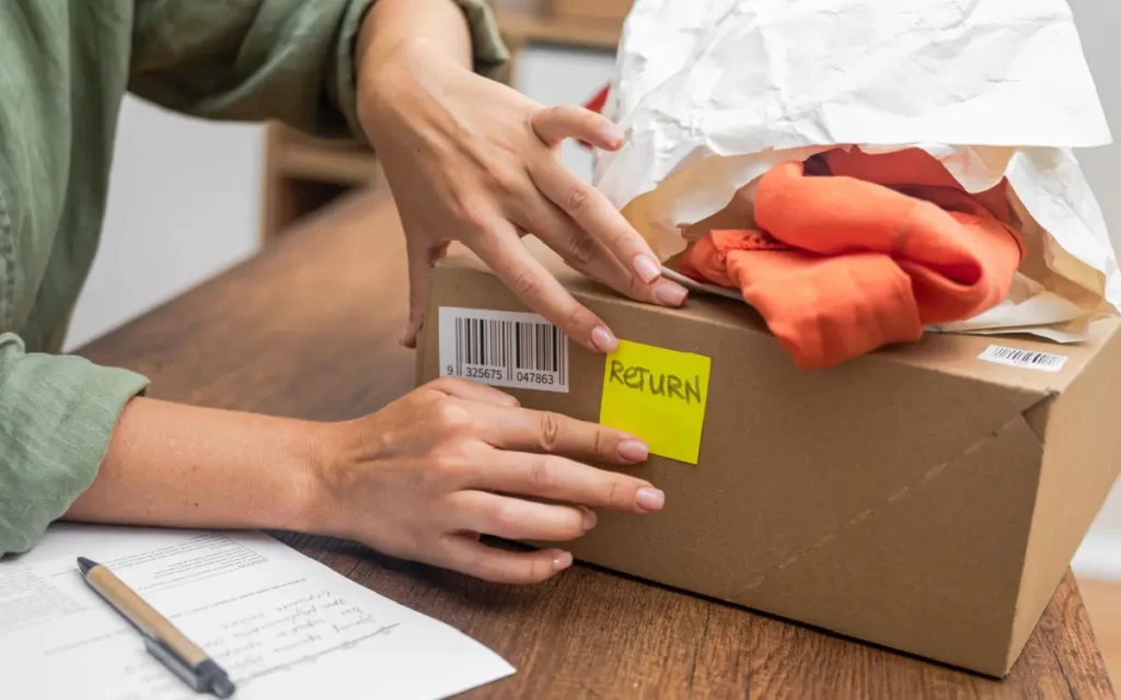 Female hands applying a yellow 'RETURN' sticker to a cardboard box with a garment in paper wrapping sitting on top of the box. Concept of online returns.