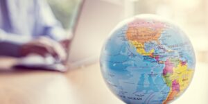 Image of a globe placed on a desk with a person sat typing on a laptop behind it.