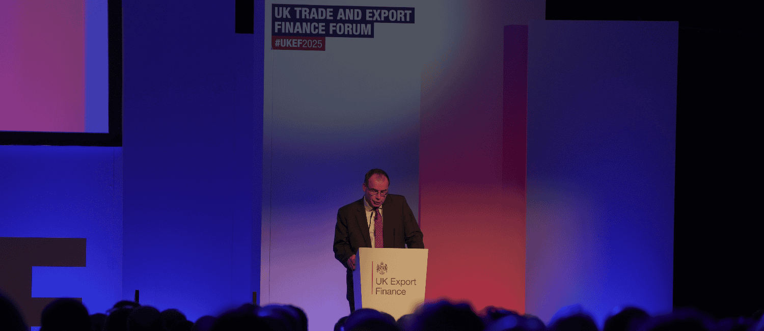 Gareth Thomas, Minister for Exports, speaks at a podium during the UK Trade and Export Finance Forum, with a backdrop displaying event branding and a blue and red-lit stage