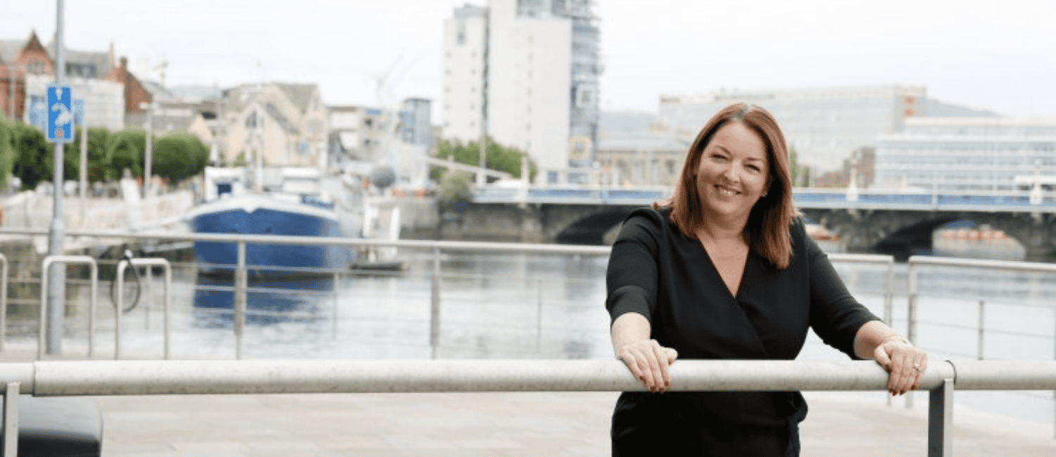 Tina McKenzie, Policy Chair at the Federation of Small Businesses, smiling in a black dress and leaning on a metal railing by a waterfront, with a cityscape, boats, and a bridge in the background