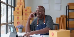 Owner of a business startup, working at his desk with boxes piled up behind him, and speaking to an insurance broker about business insurance.