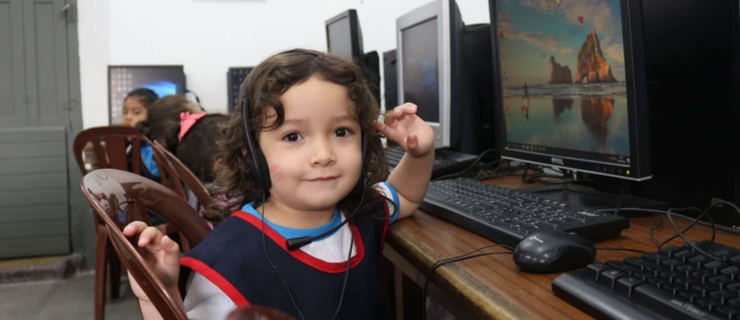 A child wearing headphones gestures playfully while seated at a computer in a classroom surrounded by other children with similar setups.