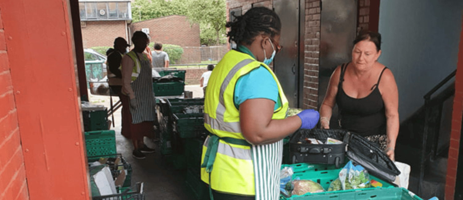 Volunteers wearing high-visibility vests and aprons are organising and distributing food at the ASTOP Divine Rescue food bank. They are surrounded by green crates filled with fresh produce and packaged goods. A woman in a black vest top is receiving items from one of the volunteers.